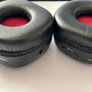 May include: Close-up of two black over-ear headphones. The earcups are black with a red interior. The headphones have volume and playback controls on the side. The earcups are made of a soft, leather-like material.