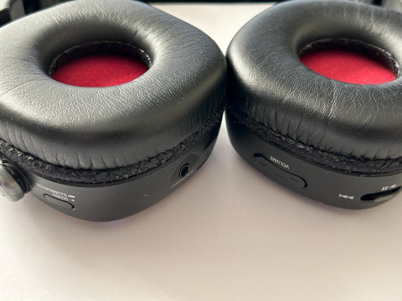May include: Close-up of two black over-ear headphones. The earcups are black with a red interior. The headphones have volume and playback controls on the side. The earcups are made of a soft, leather-like material.