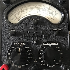 May include: A black Universal Avometer, a vintage electrical testing device. It features a central analog dial with a needle, multiple range selection knobs, and various input terminals. The device has text labels for voltage, current, and resistance measurements.