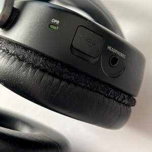 May include: Close-up of a black over-ear headphone. The headphone has a USB port, a headphone jack, and the word "HEADPHONES" printed on the side. The ear cushion is black and stitched.