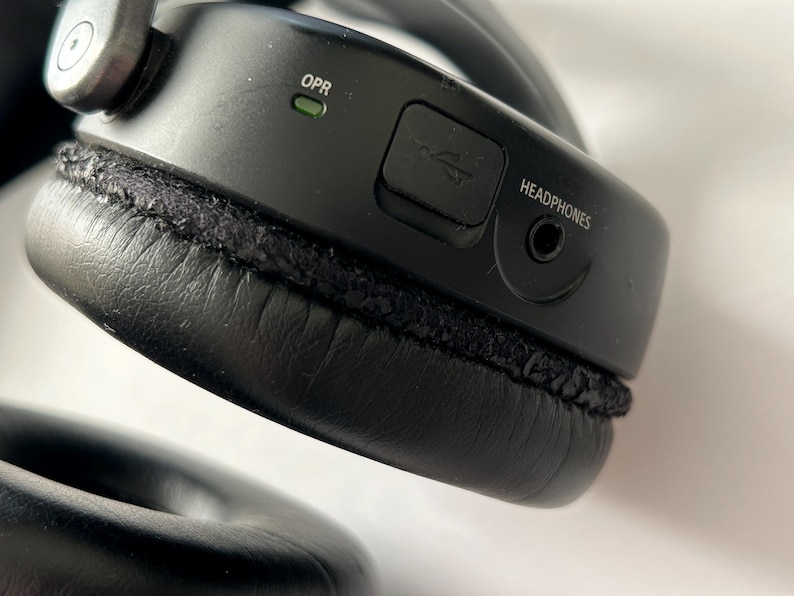 May include: Close-up of a black over-ear headphone. The headphone has a USB port, a headphone jack, and the word "HEADPHONES" printed on the side. The ear cushion is black and stitched.