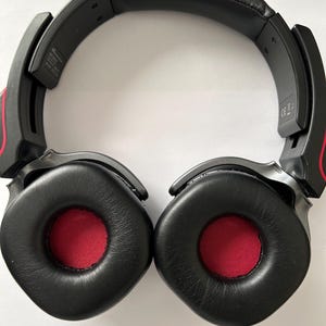 May include: Black over-ear headphones with red accents. The earcups are black with red centers. The headband is black and padded. The headphones are designed for comfort and audio quality.