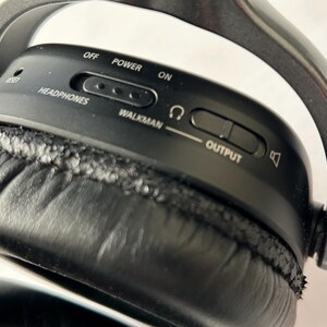 May include: Close-up of black headphones with a silver headband. The control panel has the words "OFF", "POWER", and "ON". There are also controls for "HEADPHONES", "WALKMAN", and "OUTPUT". The ear cushion is black.
