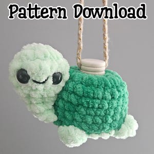 Könnte beinhalten: Eine handgefertigte Plüschschildkröte in Grüntönen, mit einem lächelnden Gesicht und großen schwarzen Augen. Die Schildkröte wird von einer beigen Schnur gehalten und trägt den Text "Pattern Download" in fetter Schrift.
