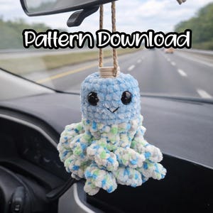 Pode incluir: Um amuleto de carro de polvo de crochê azul e branco pendurado em um espelho retrovisor. O amuleto tem olhos pretos e um rosto sorridente. O texto "Pattern Download" é exibido em letras brancas.