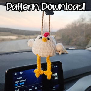 May include: A handmade white crochet chicken car charm with yellow legs and a red comb. The charm is hanging in front of a car dashboard with the text "Pattern Download" above it. The car's infotainment screen displays music information.