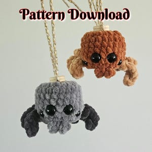 Puede incluir: Dos adornos de araña de peluche tejidos a ganchillo que cuelgan de hilos dorados y blancos, uno marrón y otro gris. Cada araña tiene grandes ojos de plástico negro y un cuerpo de hilo texturizado. El texto Pattern Download es visible arriba.