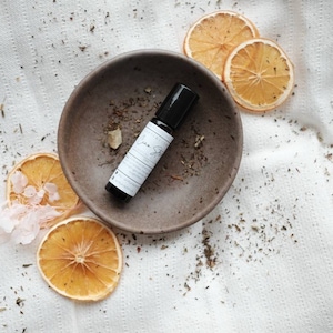 Könnte beinhalten: Eine kleine braune Keramikschale mit einer schwarzen Rollflasche mit ätherischem Öl. Die Schale befindet sich auf einem weißen Stoffhintergrund mit getrockneten Orangenscheiben.
