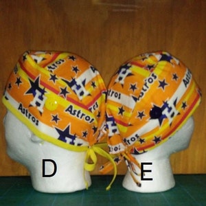 May include: Two orange, yellow, and blue patterned fabric scrub hats with the word "Astros" and a star printed on them. The hats have a button on the top and ties on the back.