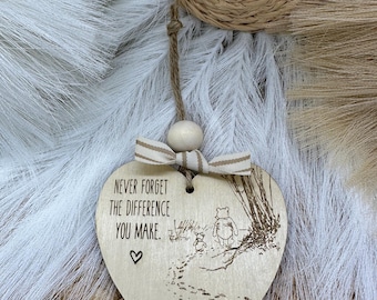 May include: Heart-shaped wooden ornament with the text "NEVER FORGET THE DIFFERENCE YOU MAKE" and a small heart symbol. The ornament features an engraved illustration of Winnie the Pooh and Piglet. A beige and white striped ribbon and wooden bead are attached.
