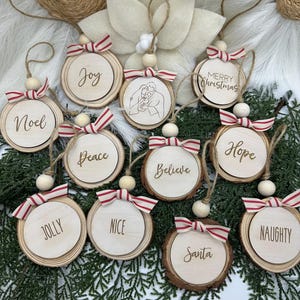 Pode incluir: Enfeites de Natal de madeira com várias palavras e frases, incluindo "Joy", "Noel" e "Merry Christmas". Cada enfeite tem um laço listrado vermelho e branco e um laço de barbante para pendurar. Os enfeites são exibidos em um leito de vegetação.