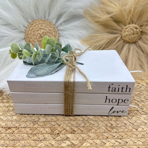 May include: Three white decorative books tied with twine, featuring the words "faith," "hope," and "love" in black script. The books are decorated with faux greenery. The background includes woven textures and floral decorations.