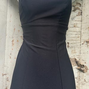 Pu&ograve; includere: Un abito nero senza maniche con scollo rotondo e un corpetto aderente caratterizzato da cuciture verticali. L'abito &egrave; abbinato a una collana composta da una sottile catenina scura e un ciondolo decorativo con tre pietre ovali viola.