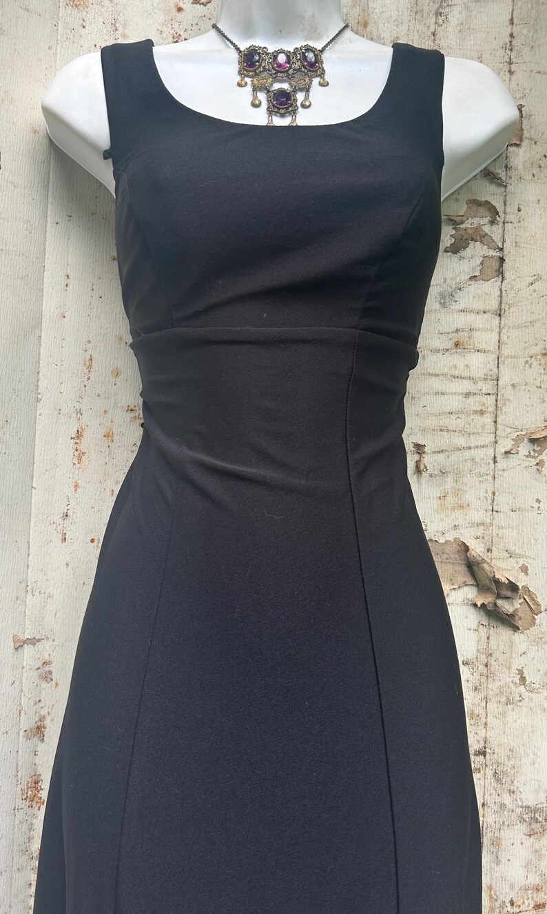 Pu&ograve; includere: Un abito nero senza maniche con scollo rotondo e un corpetto aderente caratterizzato da cuciture verticali. L'abito &egrave; abbinato a una collana composta da una sottile catenina scura e un ciondolo decorativo con tre pietre ovali viola.