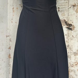Pu&ograve; includere: Un abito nero senza maniche lungo fino ai piedi con scollo rotondo e corpetto aderente caratterizzato da cuciture verticali. L'abito &egrave; abbinato a una collana color argento con catena in metallo scuro e tre pendenti decorati con pietre viola.