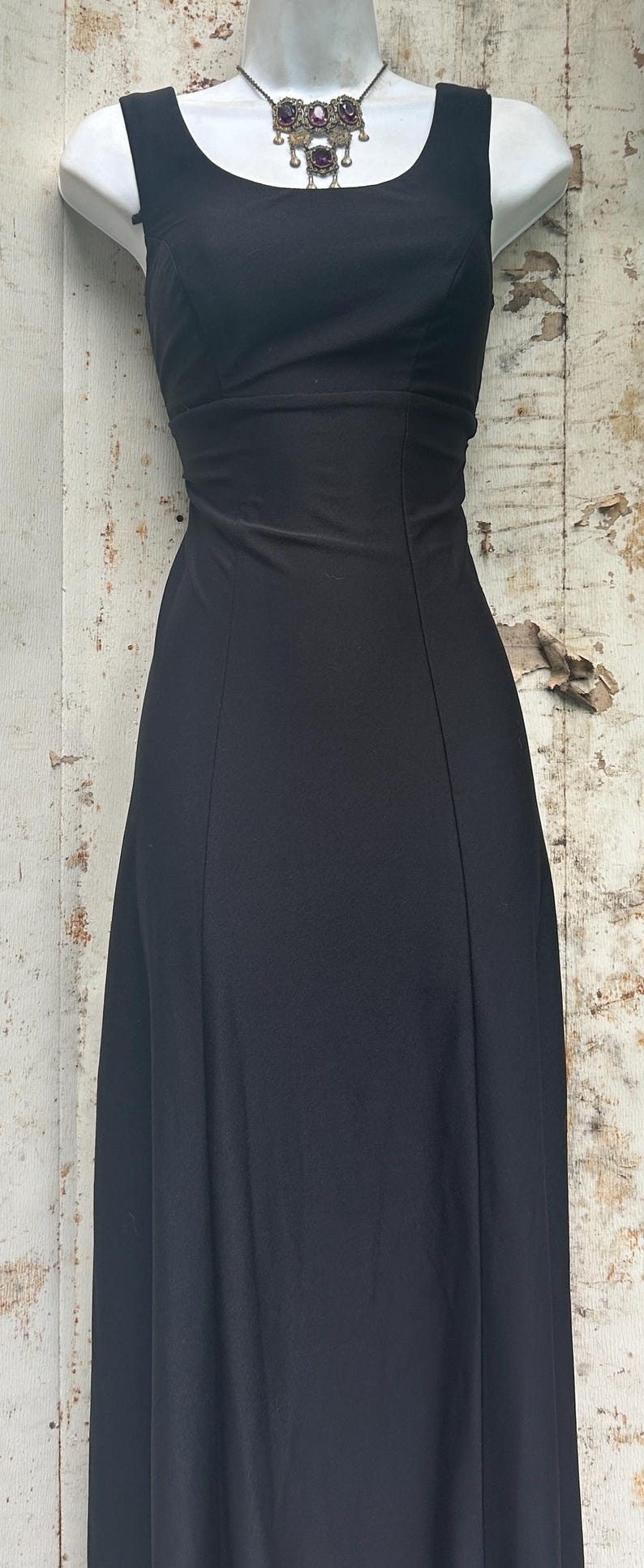 Pu&ograve; includere: Un abito nero senza maniche lungo fino ai piedi con scollo rotondo e corpetto aderente caratterizzato da cuciture verticali. L'abito &egrave; abbinato a una collana color argento con catena in metallo scuro e tre pendenti decorati con pietre viola.