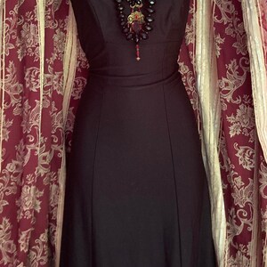 Pu&ograve; includere: Un abito nero senza maniche lungo fino ai piedi con scollo rotondo, abbinato a una collana a pi&ugrave; fili con perline sfaccettate nere e rosse e un ciondolo centrale rosso, indossato con un accessorio a colletto in pizzo bianco.