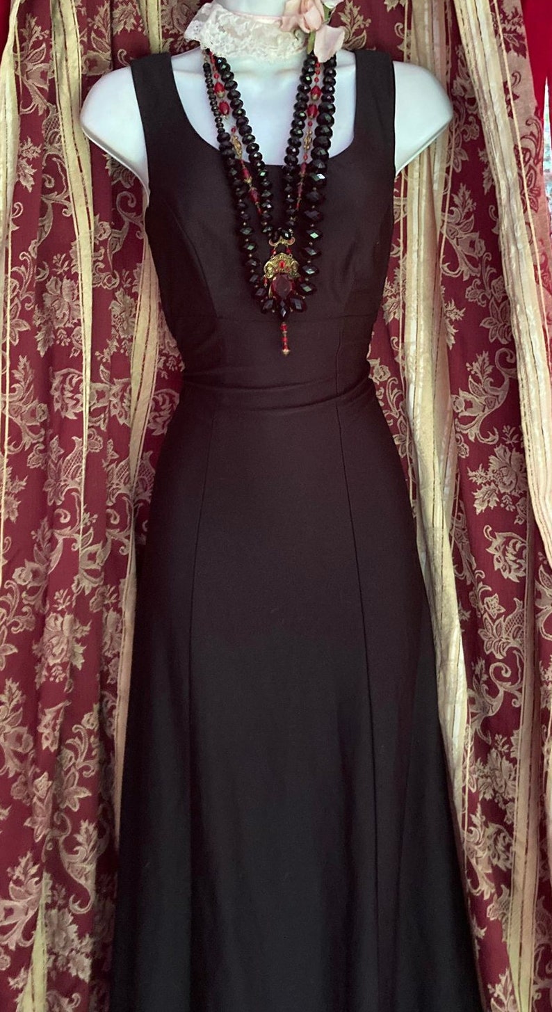 Pu&ograve; includere: Un abito nero senza maniche lungo fino ai piedi con scollo rotondo, abbinato a una collana a pi&ugrave; fili con perline sfaccettate nere e rosse e un ciondolo centrale rosso, indossato con un accessorio a colletto in pizzo bianco.