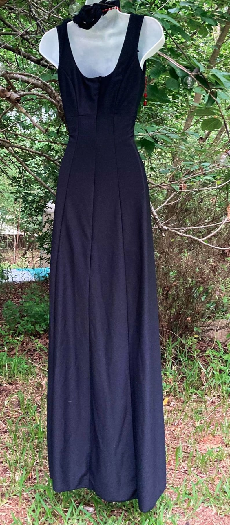 Pu&ograve; includere: Un abito nero senza maniche lungo fino ai piedi con scollo rotondo, un corpetto aderente con cuciture verticali e una gonna lunga e fluida. Il capo &egrave; esposto su un manichino bianco, mostrando un design in tinta unita e una silhouette semplice.