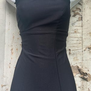 Pu&ograve; includere: Un abito nero senza maniche con scollo rotondo e corpetto aderente caratterizzato da cuciture verticali. L'abito &egrave; abbinato a una collana importante composta da una catena in metallo scuro e diversi pendenti decorati con pietre ovali viola.