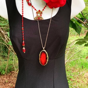 Pu&ograve; includere: Un abito nero senza maniche abbinato a una grande rosa rossa, un girocollo in pizzo nero e due collane sovrapposte. Una collana presenta perline rosse, mentre l'altra mostra un grande ciondolo ovale rosso incastonato in una cornice dorata.