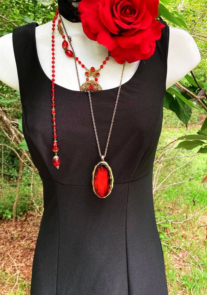 Pu&ograve; includere: Un abito nero senza maniche abbinato a una grande rosa rossa, un girocollo in pizzo nero e due collane sovrapposte. Una collana presenta perline rosse, mentre l'altra mostra un grande ciondolo ovale rosso incastonato in una cornice dorata.