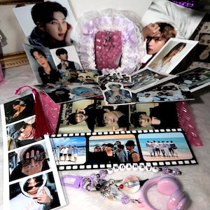 May include: A collection of K-pop merchandise including multiple photographic prints of band members, a film strip style photo card, a pink ruffled frame, and a beaded keychain with a purple clasp and a pink headphone charm featuring the name TAEHYUNG.