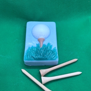 May include: A rectangular, light blue soap bar with a golf ball design, a brown tee, and green grass. Three wooden golf tees are scattered below the soap bar. The background is a solid green color.