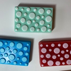 May include: Three rectangular bars of soap with a polka dot pattern. The soap on the top is light green with white polka dots. The soap on the left is blue with light blue polka dots. The soap on the right is red with pink polka dots.