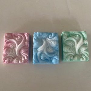 May include: Three square-shaped bars of soap with a swirling white design on a pink, blue, and green background.