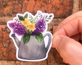 Puede incluir: Una ilustración de acuarela de una regadera gris llena de flores moradas y amarillas. Las flores están pintadas en un estilo realista y la regadera tiene un fondo blanco.