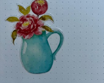 May include: A blue ceramic pitcher with a single handle, filled with pink and red flowers. The pitcher is decorated with a watercolor-style design.