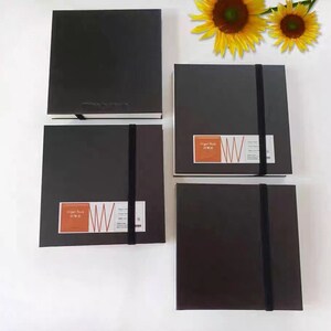 May include: Four black sketchbooks with elastic closures. Each sketchbook has a white label with the text "Sketch Book" and a red and white logo. The sketchbooks are arranged in a row on a white surface.