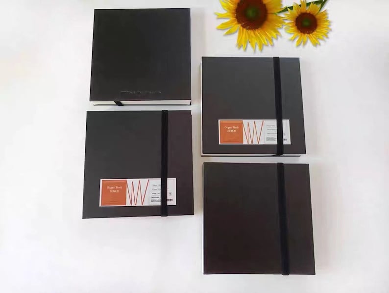 May include: Four black sketchbooks with elastic closures. Each sketchbook has a white label with the text "Sketch Book" and a red and white logo. The sketchbooks are arranged in a row on a white surface.
