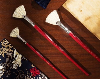 May include: Three fan-shaped paint brushes with red wooden handles and silver metal ferrules. The brushes are on a wooden surface with a piece of fabric and a piece of paper in the background.
