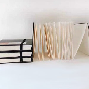 May include: Two stacks of black notebooks with black elastic bands. One stack is closed and the other is open, revealing blank white pages.