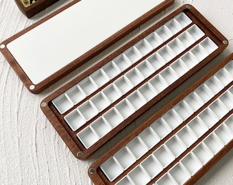 May include: A rectangular wooden watercolour palette box with a white mixing surface and two hinged sections containing multiple small white square paint wells. A small rectangular wooden block with the text AAAI and a gold metal clip are included.