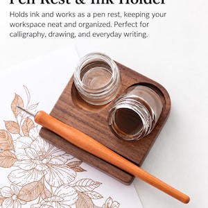 May include: A dark wood pen rest and ink holder featuring two circular cutouts containing clear glass jars. A light brown wooden pen rests horizontally across the base. Text at the top reads: Pen Rest & Ink Holder. Holds ink and works as a pen rest, keeping your workspace neat and organised. Perfect for calligraphy, drawing, and everyday writing.