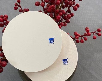 May include: Two off-white, round coasters with a blue logo, stacked on top of each other. The coasters are next to a sprig of red berries. The logo on the coasters reads "BACHONG".