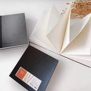 May include: Two black sketchbooks with a textured cover. One sketchbook is closed and the other is open to reveal white paper. The open sketchbook has the text "Sketch Book 150 gsm" printed on the cover.