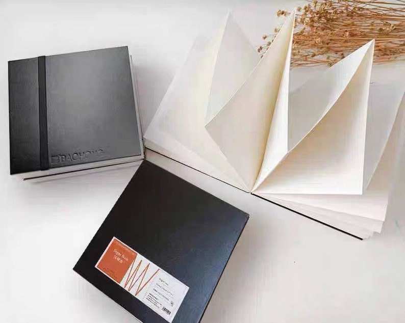 May include: Two black sketchbooks with a textured cover. One sketchbook is closed and the other is open to reveal white paper. The open sketchbook has the text "Sketch Book 150 gsm" printed on the cover.