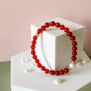 May include: A red beaded bracelet with gold accents and a single white pearl bead. The bracelet is displayed on a white geometric surface with scattered white pearls, set against a pink and white background. The beads are round and evenly spaced.