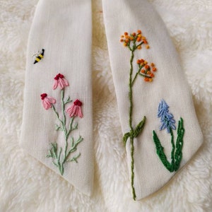 May include: A white fabric bow tie with embroidered flowers and a bee. The flowers are pink, orange, and blue.