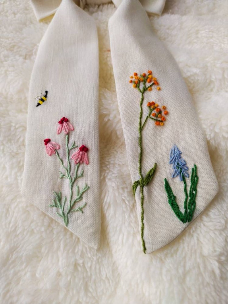 May include: A white fabric bow tie with embroidered flowers and a bee. The flowers are pink, orange, and blue.