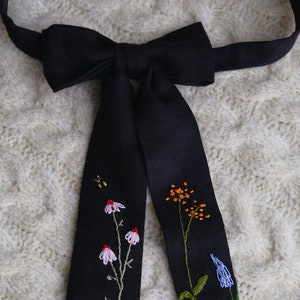 May include: A black bow tie featuring embroidered flowers in shades of pink, orange, green, and blue. The bow tie is crafted from a soft, textured fabric.