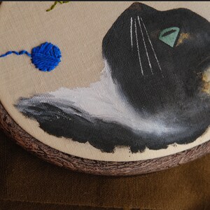 May include: Embroidered hoop art featuring a black and white cat with a green eye, looking up at a blue yarn ball. The hoop is made of wood with a brown finish.