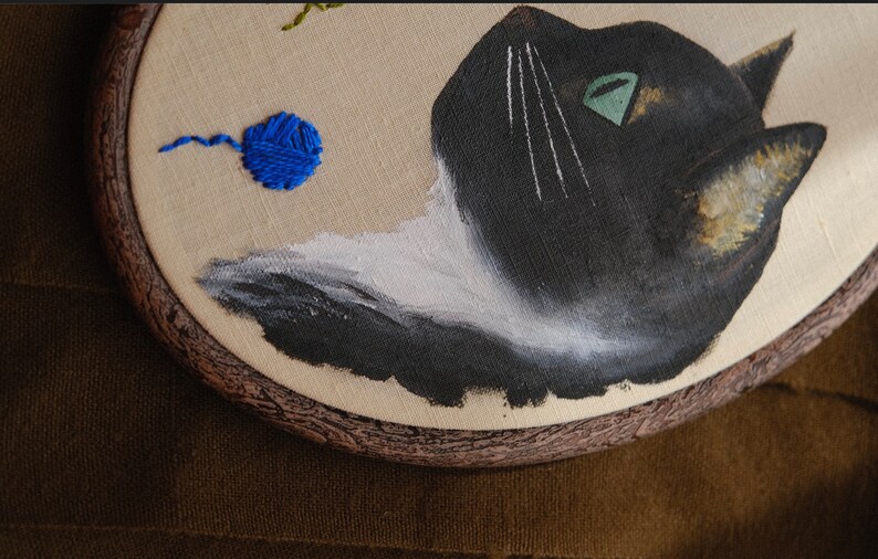 May include: Embroidered hoop art featuring a black and white cat with a green eye, looking up at a blue yarn ball. The hoop is made of wood with a brown finish.