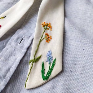 May include: A white fabric tie with embroidered flowers in shades of green, orange, and blue. The tie is on a light blue fabric background.