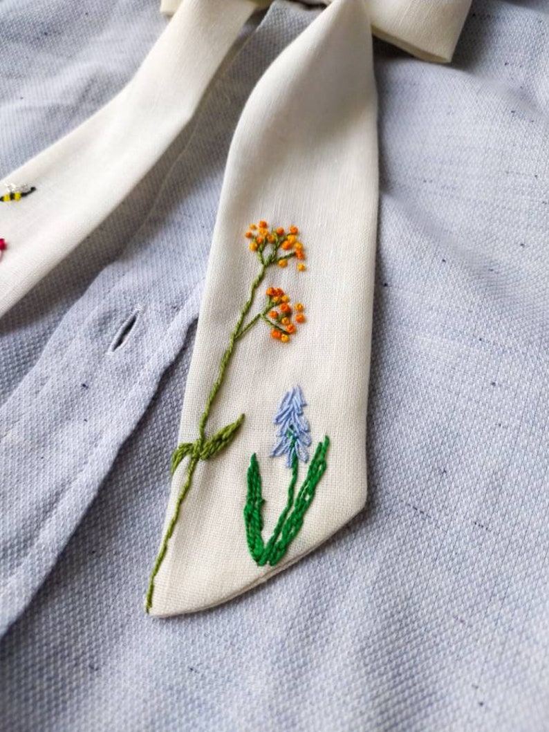 May include: A white fabric tie with embroidered flowers in shades of green, orange, and blue. The tie is on a light blue fabric background.