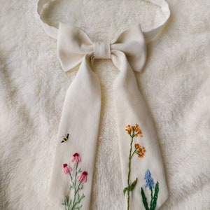 May include: A white linen bow tie with embroidered flowers and a bee. The flowers are pink, orange, blue, and green.